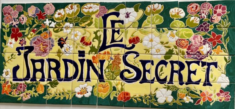 Plaque « Le Jardin Secret » : lettres bleu nuit sur fond jaune entouré de fleurs, carreaux de céramique peints à la main