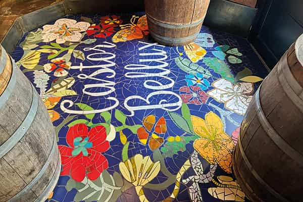 Mosaïque Castor Bellux posée dans l'entrée du restaurant à Dinard, entourée de tonneaux en bois, lettrage cursif blanc et fleurs colorées
