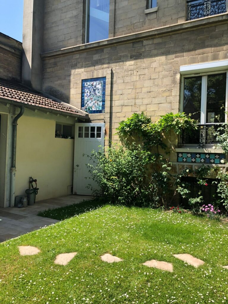 Façade en pierre côté jardin : sous une fenêtre à garde-corps en fer forgé, une frise de carreaux bleus et turquoise à fleurs blanches en relief, et un panneau bleu dans une niche en hauteur