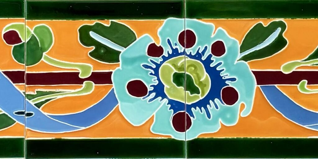 Détail de la frise d'entrée de Colombes : une fleur turquoise et un ruban bleu sur fond orange, entre deux bandes vert sombre