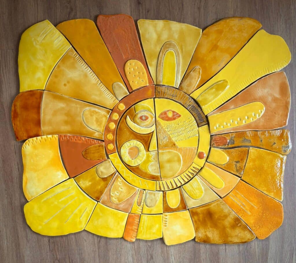Soleil en céramique posé à plat sur un parquet : rayons de pièces découpées jaunes, ocres, orangées et brun rouille autour d'un visage