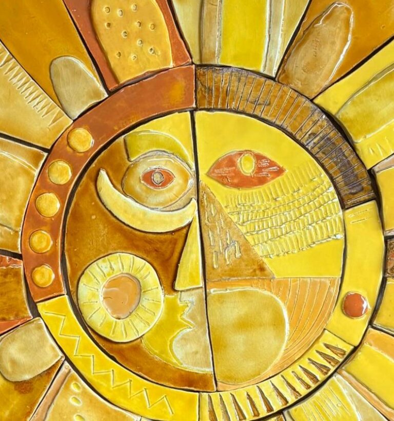 Le visage du soleil en céramique : à gauche un œil rond et un croissant sur fond brun, à droite un œil en amande et un nez en triangle sur fond jaune
